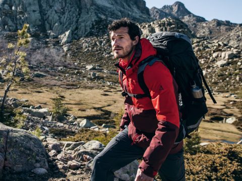 Eine Person in einer roten Jacke wandert mit einem großen Rucksack durch eine Bergregion. Die Landschaft ist von felsigem Gelände und spärlicher Vegetation unter einem klaren blauen Himmel geprägt.