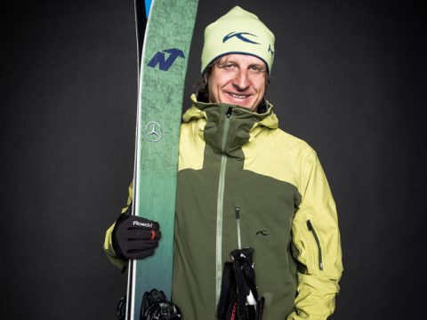 Eine Person mit grüner Skijacke und Mütze lächelt und hält dabei die Skier hoch. Die Jacke hat eine hellgrüne Kapuze und verschiedene Logos, darunter ein Mercedes-Emblem. Der Hintergrund ist dunkel und hebt das Motiv hervor.