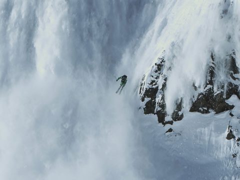 Ein Skifahrer in einer grünen Jacke fährt eine extreme Abfahrt eine schneebedeckte Klippe hinunter, umgeben von Schnee und Nebel. Die dynamische Bewegung fängt den Nervenkitzel des Skifahrens in einer herausfordernden Bergumgebung ein.