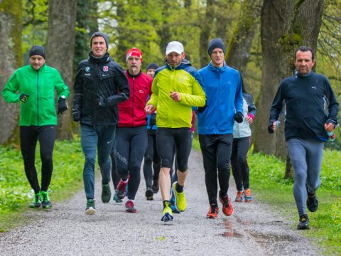 Eine Gruppe von Menschen joggt gemeinsam auf einem von Bäumen gesäumten Weg. Sie tragen farbenfrohe Jacken und Laufkleidung, die von Rot und Grün bis hin zu Blau und Schwarz reicht. Die Szenerie ist im Freien, mit Gras und Bäumen, was an einen Park oder einen Naturpfad erinnert.