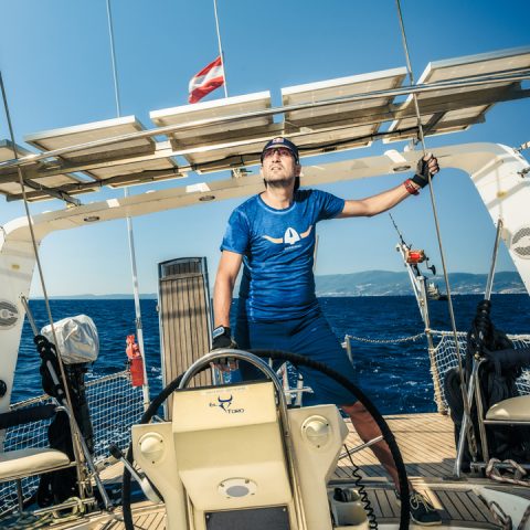 Eine Person in blauem Hemd und Mütze steht selbstbewusst auf dem Deck eines Segelboots und hält ein Seil. Im Hintergrund ist der Ozean mit einem klaren blauen Himmel zu sehen, und oben ist eine Flagge zu sehen. Im Vordergrund ist das Steuerrad zu sehen.