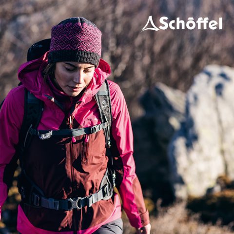 Eine Person in einer rot-rosa Wanderjacke und Strickmütze wandert im Freien. Sie trägt einen Rucksack. Der Hintergrund ist verschwommen und zeigt Felsen und Blätter. In der oberen rechten Ecke ist das Schöffel-Logo zu sehen.
