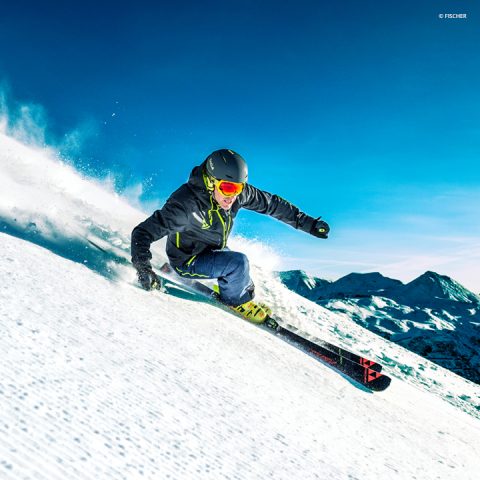 Ein Skifahrer in dunkler Jacke und Neonbrille fährt einen schneebedeckten Berghang hinunter und wirbelt dabei eine Schneewolke auf. Die Sonne scheint hell am klaren blauen Himmel, im Hintergrund sind in der Ferne schneebedeckte Gipfel zu sehen.