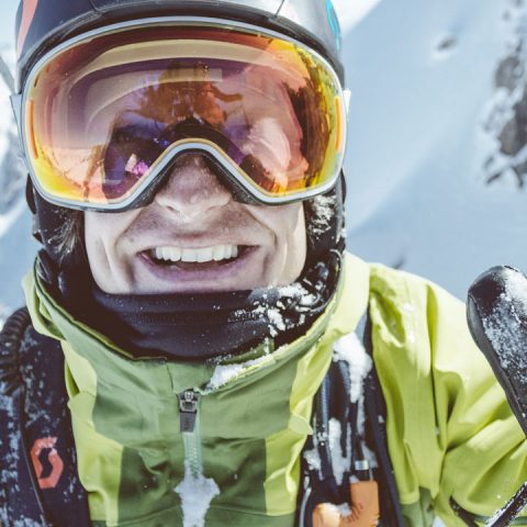 Eine Person in grüner Jacke und Skibrille steht lächelnd im Schnee. Sie scheint Ski oder Snowboard zu fahren, im Hintergrund sind schneebedeckte Berge zu sehen.