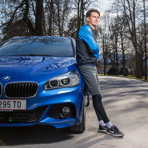 Eine Person in Sportkleidung lehnt an einem blauen BMW, der auf einer von Bäumen gesäumten Straße geparkt ist. Die Person blickt mit verschränkten Armen in die Kamera. Die Szene spielt an einem klaren Tag und das Nummernschild ist teilweise sichtbar.