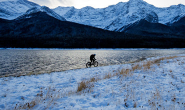 Ein Radfahrer fährt auf einem schneebedeckten Weg neben einem großen Gewässer entlang, im Hintergrund sind schneebedeckte Berge unter einem leicht bewölkten Himmel zu sehen.