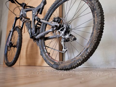 Eine Nahaufnahme eines schlammigen Mountainbikes, das drinnen auf einem Holzboden abgestellt ist. Um die Reifen herum ist Schmutz verstreut. Das Fahrrad ist schräg gestellt, sodass die Federung und die Gangschaltung sichtbar sind.