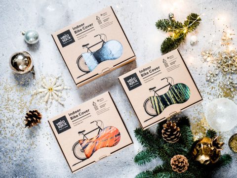 Drei verpackte Velosock-Fahrradabdeckungen für den Innenbereich in blauen, grünen und orangefarbenen Mustern sind auf einem festlichen Hintergrund mit Tannenzweigen, Ornamenten und goldenem Konfetti angeordnet.