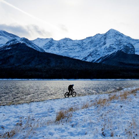 Silhouette einer Person, die auf einem verschneiten Weg neben einem See Fahrrad fährt. Im Hintergrund erheben sich schneebedeckte Berge unter einem bewölkten Himmel. Die Landschaft ist ruhig und die Berge spiegeln sich auf der Wasseroberfläche.