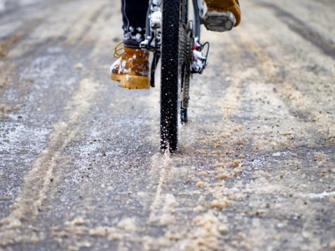 Nahaufnahme eines Fahrradreifens auf einer matschigen, schneebedeckten Straße. Der Fahrer trägt hellbraune Stiefel und der Reifen verdrängt den Matsch. Das Bild fängt die Textur und Bewegung des Fahrradfahrens durch einen winterlichen Mix ein.