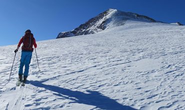 Eine Person in roter Jacke und blauer Hose fährt unter einem klaren blauen Himmel einen schneebedeckten Hang hinauf auf einen Berggipfel. Auf dem Schnee sind Skispuren und lange Schatten zu sehen.