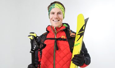 Eine lächelnde Person trägt eine rote Steppjacke, ein grünes Stirnband und hält leuchtend gelbe Skier und Skistöcke in der Hand. Sie hat einen Rucksack dabei und steht vor einem schlichten weißen Hintergrund.