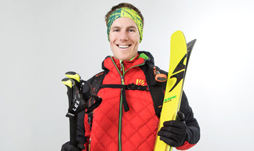 Eine lächelnde Person trägt eine rote Steppjacke, ein grünes Stirnband und hält leuchtend gelbe Skier und Skistöcke in der Hand. Sie hat einen Rucksack dabei und steht vor einem schlichten weißen Hintergrund.