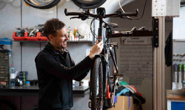 Eine Person in einer Werkstatt stellt ein schwarzes Fahrrad auf einem Reparaturständer ein. Im Hintergrund sind Werkzeuge, Regale und Fahrradräder an der Wand montiert. Die Person lächelt und ist auf die Aufgabe konzentriert.