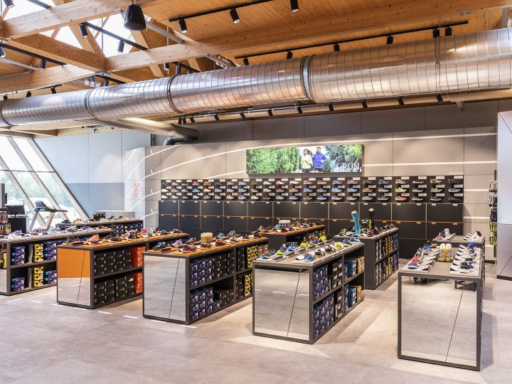 Ein modernes Schuhgeschäft mit Holzdecken, freiliegenden Leitungen und großen Fenstern. Schuhdisplays mit verschiedenen Schuhstilen sind im ganzen Raum verteilt. Ein digitaler Bildschirm an der Rückwand zeigt ein Bild von Menschen im Freien.