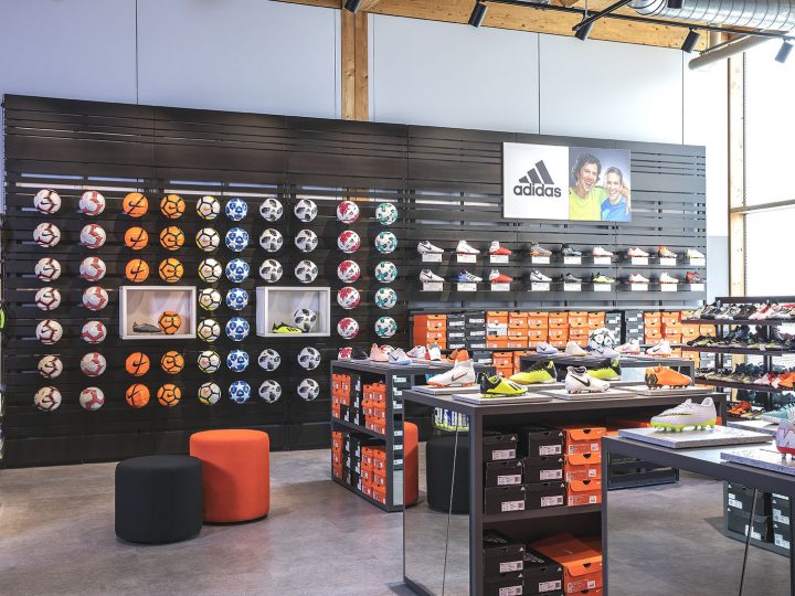 Inneneinrichtung eines Sportgeschäfts mit einer Wandpräsentation verschiedener Fußbälle und Regalen mit Adidas-Schuhen. Darunter sind ordentlich leuchtend orangefarbene Schuhkartons gestapelt. Das Geschäft hat ein modernes Design mit Holzakzenten und Industriebeleuchtung.