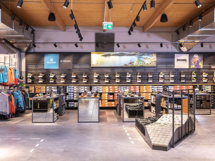 Ein modernes Schuhgeschäft mit einer Auswahl an Outdoor-Schuhen in Regalen. Marken sind auf einer Wandanzeige sichtbar. Das Geschäft verfügt über elegante Beleuchtung, verspiegelte Armaturen und einen Bereich zum Anprobieren von Schuhen auf einer strukturierten Rampe. Kleidung befindet sich auf der linken Seite.