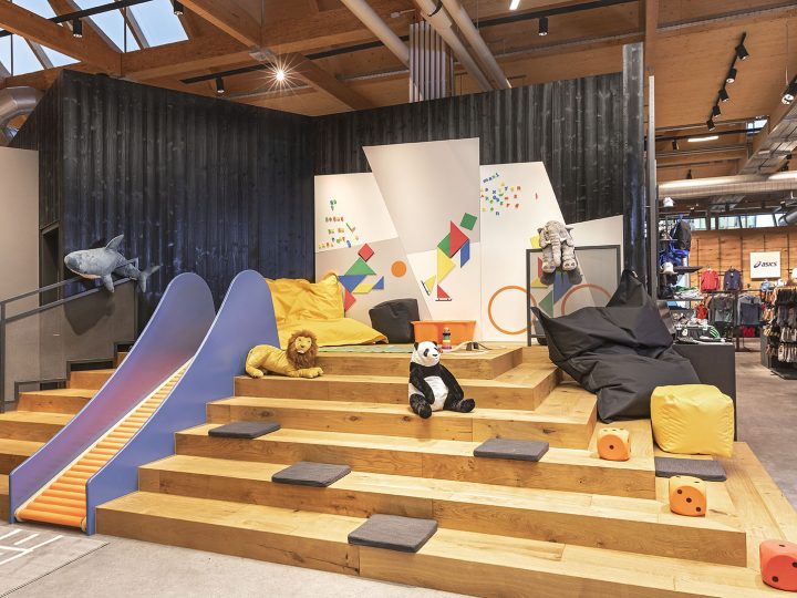 Kinderspielplatz mit Holzstufen, einer blauen Rutsche und farbenfrohen geometrischen Wanddesigns. Auf den Stufen stehen ausgestopfte Tiere, darunter ein Löwe und ein Panda, sowie Sitzsäcke. Der Raum befindet sich in einem modernen Industrieambiente mit sichtbaren Rohren und Holzakzenten.