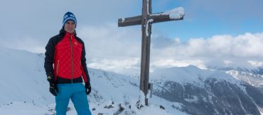 Eine Person in einer rot-schwarzen Jacke und blauen Hosen steht auf einem schneebedeckten Berg neben einem großen Holzkreuz. Im Hintergrund sind schneebedeckte Gipfel und ein teilweise bewölkter Himmel zu sehen.
