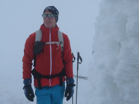 Eine Person mit einer roten Jacke, blauen Hosen, Handschuhen und Mütze steht in einer nebligen, verschneiten Landschaft. Sie hat einen Rucksack und Skistöcke dabei und steht neben einem großen, mit Frost bedeckten Gebäude.