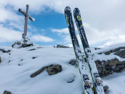 Zwei Skier stehen aufrecht im verschneiten Gelände neben einem Holzkreuz auf einem kleinen Hügel unter einem teilweise bewölkten Himmel. Schneebedeckte Felsen sind sichtbar und bilden einen Kontrast zum strahlend blauen Himmel.