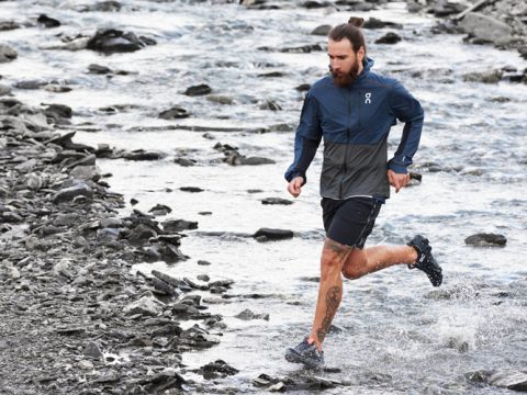 Ein Mann mit Bart und einer Tätowierung am Bein läuft über einen steinigen Bach. Er trägt eine blau-graue Jacke und schwarze Shorts. Das Wasser plätschert unter seinen Wanderschuhen, während er sich energisch durch die Natur bewegt.