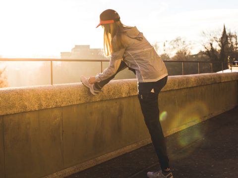 Eine Person in leichter Jacke und Mütze streckt bei Sonnenaufgang oder Sonnenuntergang ihr Bein auf einem Felsvorsprung aus. Sie befindet sich im Freien auf einem Pfad mit einer malerischen, sonnenbeschienenen Kulisse aus Bäumen und entfernten Gebäuden.