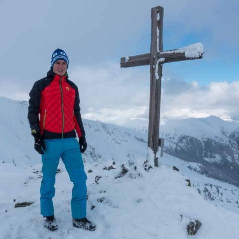 Eine Person in Winterkleidung steht neben einem Holzkreuz auf einem schneebedeckten Berggipfel. Die Person trägt eine rote Jacke, blaue Hosen und eine blaue Wintermütze. Im Hintergrund sind schneebedeckte Berge unter einem bewölkten Himmel zu sehen.