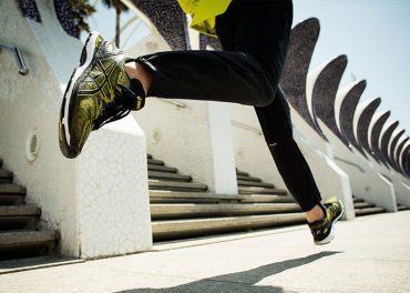 Eine Person in schwarzen Hosen und limettengrünen Turnschuhen wird mitten beim Laufen im Freien aufgenommen. Im Hintergrund sind moderne architektonische Strukturen und Steintreppen unter einem klaren Himmel zu sehen.