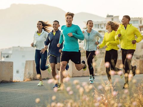 Eine Gruppe von sechs Personen joggt an einem sonnigen Tag im Freien. Sie lächeln und tragen sportliche Kleidung, darunter Jacken und Shorts. Im Hintergrund sind eine Berglandschaft und Gebäude zu sehen. Im Vordergrund sind Wildblumen zu sehen.