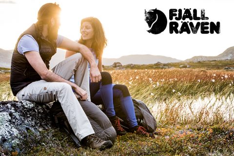 Ein Mann und eine Frau sitzen bei Sonnenuntergang in Wanderkleidung auf Felsen auf einem offenen Feld. Sie lächeln sich an und haben Rucksäcke in der Nähe. Das Fjällräven-Logo befindet sich in der oberen rechten Ecke. Im Hintergrund sind sanfte Hügel zu sehen.