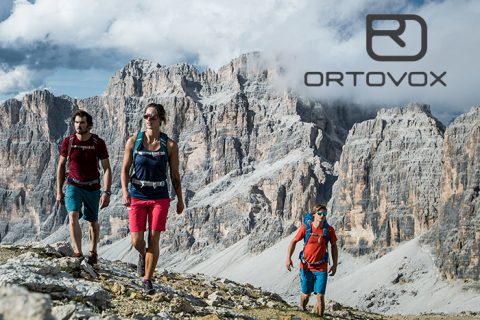 Drei Wanderer mit Rucksäcken laufen auf einem felsigen Pfad vor einer Berglandschaft unter einem bewölkten Himmel. Oben rechts ist das ORTOVOX-Logo zu sehen.