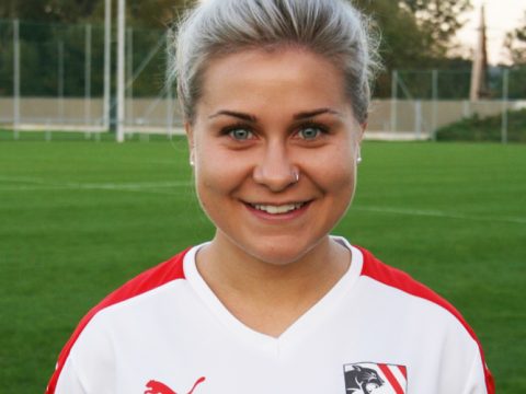 Eine Person mit kurzen blonden Haaren lächelt und trägt ein weiß-rotes Sporttrikot mit einem Logo. Sie steht auf einer Wiese mit Sportgeräten im Hintergrund.