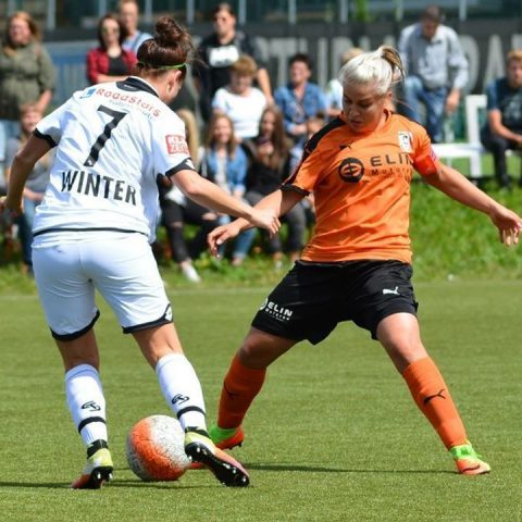 Zwei Fußballerinnen kämpfen auf einem Rasenfeld um den Ball. Die Spielerin in Weiß, Nummer 7, steht der Spielerin in Orange gegenüber. Im Hintergrund sind Zuschauer zu sehen, die das Spiel aufmerksam verfolgen.