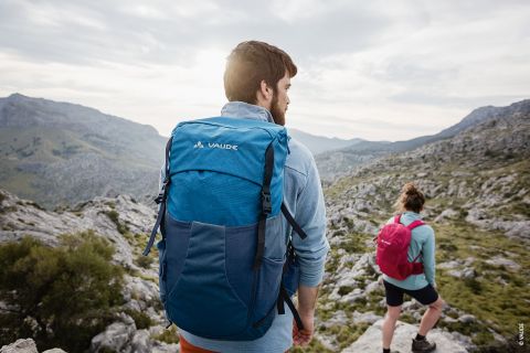 Eine Person mit einem blauen Rucksack steht auf einem felsigen Bergpfad und blickt in die Ferne. Eine andere Person mit einem rosa Rucksack sitzt auf einem Felsen in der Nähe. Die Landschaft besteht aus sanften Hügeln unter einem bewölkten Himmel.