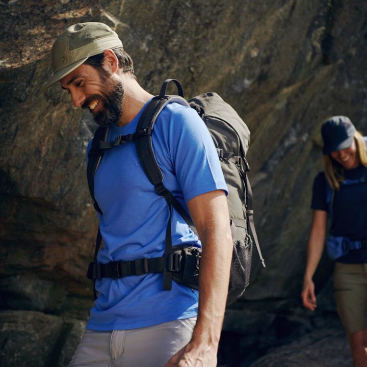 Zwei Wanderer, ein Mann und eine Frau, gehen einen steinigen Pfad unter einem großen überhängenden Felsen entlang. Der Mann vorn trägt ein blaues Hemd und einen Rucksack und lächelt. Die Frau folgt ihm, ebenfalls mit einem Rucksack, während das Sonnenlicht durch die Felsen fällt.