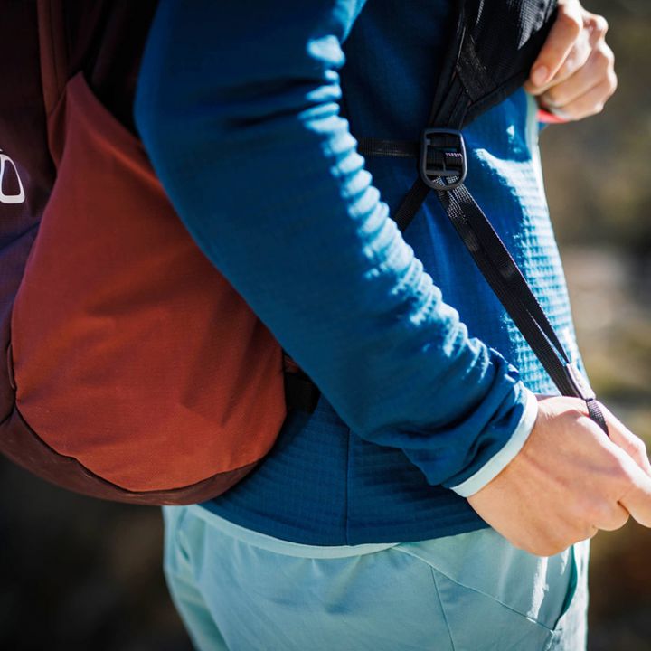 Eine Person in einem blauen Langarmoberteil und blaugrünen Hosen zieht den Riemen eines braunen Rucksacks fest. Im Hintergrund ist eine verschwommene Naturlandschaft zu sehen, die eine Szenerie im Freien suggeriert.