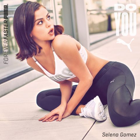 Eine Frau in Sportkleidung sitzt auf einem Betonboden neben einem Fenster und macht Dehnübungen. Sie trägt einen weißen Sport-BH und schwarze Leggings. Im Hintergrund sind die Worte „FOREVER FASTER PUMA“ und „DO YOU“ zu sehen.