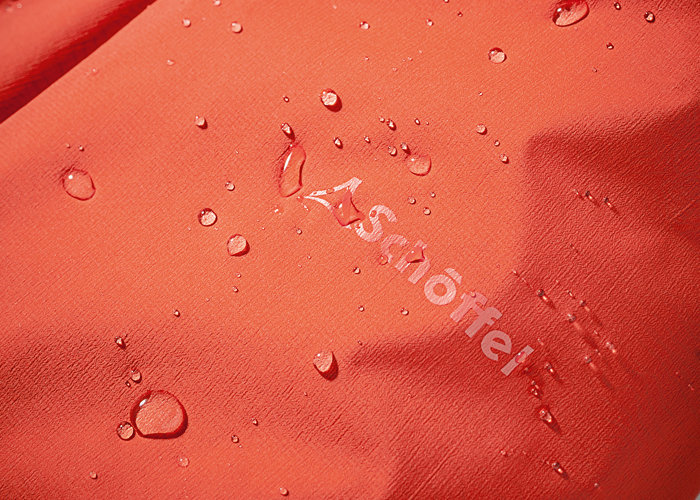 Nahaufnahme von Wassertropfen, die auf einer glatten, orangefarbenen Stoffoberfläche abperlen. Das Wort „Schöffel“ ist teilweise sichtbar.