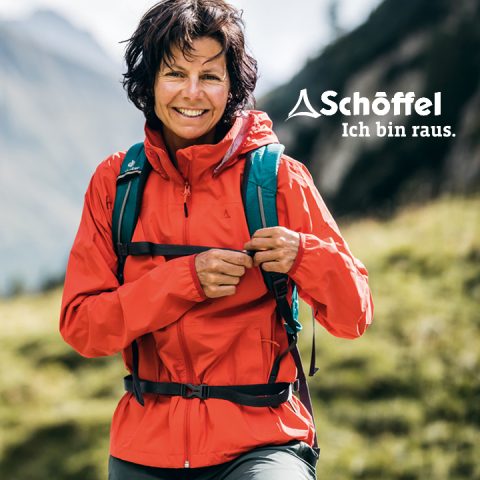 Eine Person steht lächelnd mit roter Outdoorjacke und Rucksack in einer Berglandschaft. Zu sehen sind das Schöffel-Logo und der Schriftzug „Ich bin raus.“.