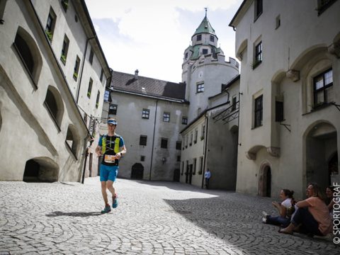 Ein Läufer in Sportkleidung joggt durch einen gepflasterten Innenhof, der von historischen Gebäuden mit Bogenfenstern umgeben ist. Im Hintergrund ist ein hoher Turm zu sehen. Zwei Personen sitzen auf dem Boden und beobachten den Läufer. Der Himmel ist teilweise bewölkt.