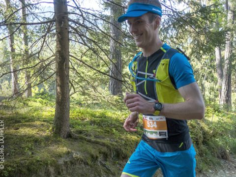 Eine Person läuft im Trailrunning durch einen Wald. Sie trägt ein blau-gelbes Sportoutfit, eine blaue Mütze und die Startnummer 534. Der Hintergrund ist voller Bäume und Grün.