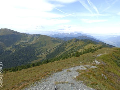 Eine malerische Aussicht auf eine Berglandschaft unter einem teilweise bewölkten Himmel. Sanfte Hügel, bedeckt mit grünem Gras und Baumgruppen, erstrecken sich in die Ferne. Im Vordergrund ist ein schmaler, gewundener Pfad zu sehen.