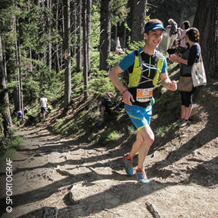 Ein lächelnder Läufer in Sportkleidung und mit Visier joggt während eines Rennens einen Waldweg bergauf. Zuschauer stehen in der Nähe und Sonnenlicht fällt durch die Bäume.