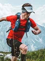 Eine Person mit rotem Shirt, schwarzen Shorts und blauer Laufweste läuft in einer Bergregion Trailrunning. Sie trägt ein Visier und ein Stirnband und wirkt konzentriert, vor der Kulisse von Wolken und schroffen Gipfeln. Im Vordergrund sind Gras und Blumen zu sehen.