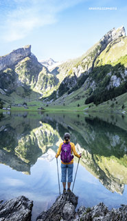 Eine Person mit einem lila Rucksack und Wanderstöcken steht auf Felsen neben einem ruhigen See, im Wasser spiegeln sich Berge und ein klarer Himmel, umgeben von üppigem Grün.