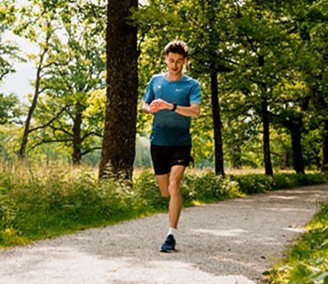 Eine Person in blauem Hemd und schwarzen Shorts läuft auf einem Kiesweg in einem üppigen, grünen Park. Sie überprüft ein Gerät an ihrem Handgelenk. Sonnenlicht fällt durch die Bäume und erzeugt gesprenkelte Schatten auf dem Boden.