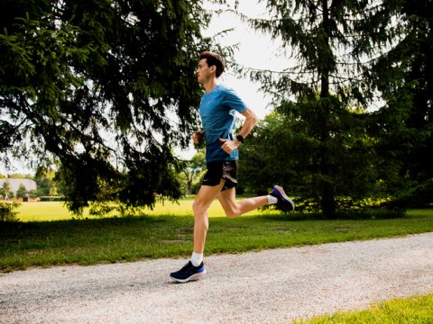 Eine Person in blauem Hemd und schwarzen Shorts joggt auf einem Schotterweg durch einen üppig grünen Park. Hohe Bäume säumen den Weg und Sonnenlicht fällt durch die Äste und schafft eine ruhige Außenatmosphäre.