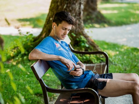 Ein Mann in einem blauen Hemd sitzt auf einer Parkbank, hört über Kopfhörer Musik und schaut auf sein Smartphone. Er steht an einem sonnigen Tag unter einem Baum, umgeben von Grün.