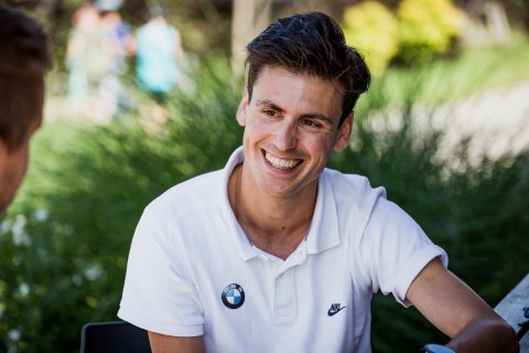 Lächelnder Mann in weißem Poloshirt mit Markenlogos sitzt draußen und unterhält sich. Im Hintergrund sind verschwommenes Grün und Menschen zu sehen.
